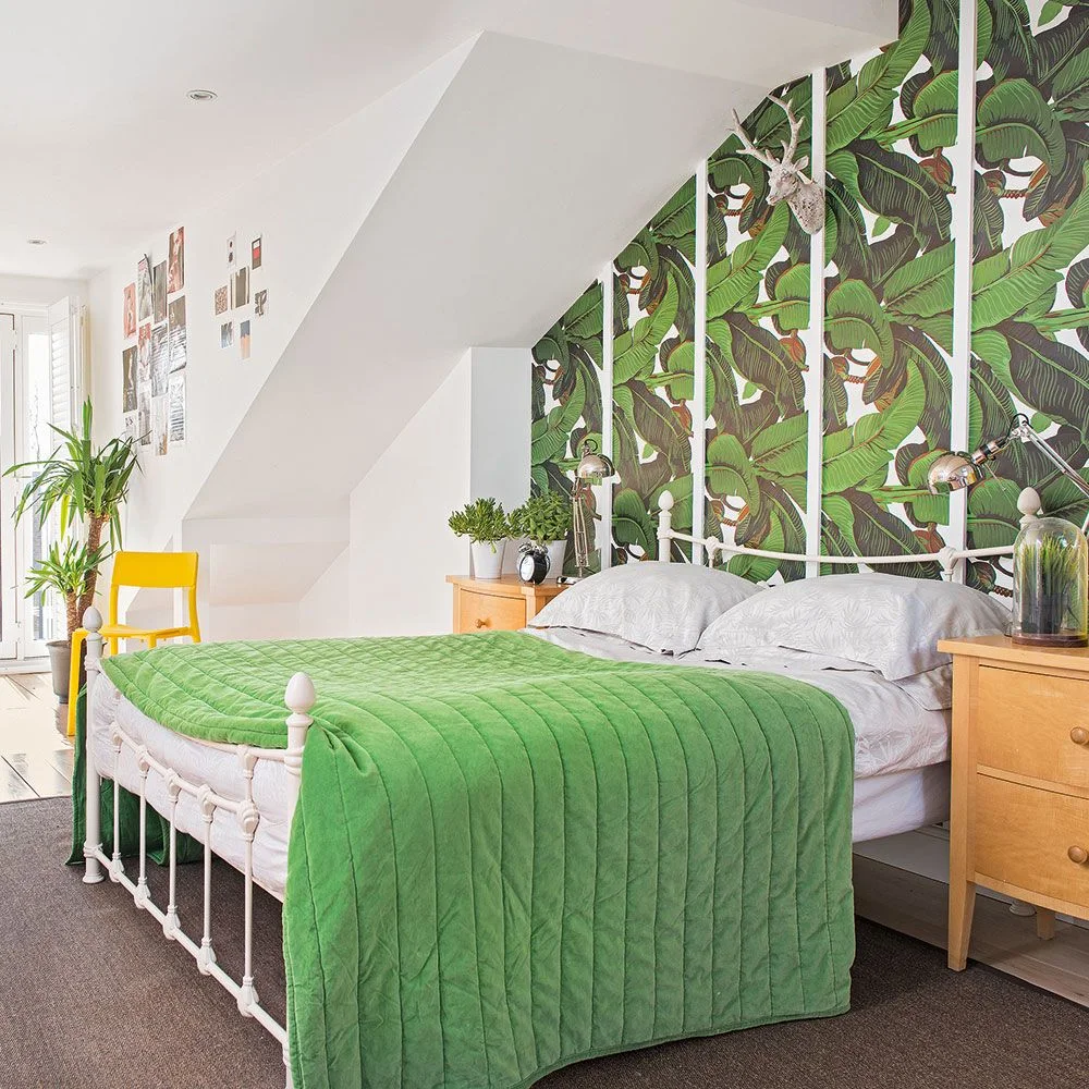 A white metal bed with a bright green quilt sits beneath a large leaf-patterned wall panel beside wooden drawers and a yellow chair
