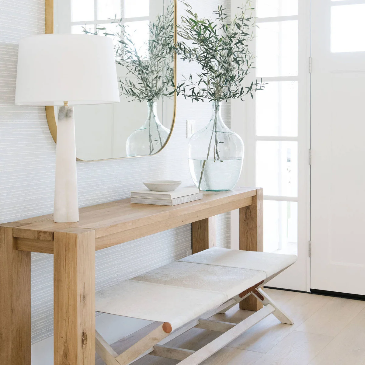 A pale wood console with a tall white lamp and glass vase, with two light folding-style stools tucked underneath.