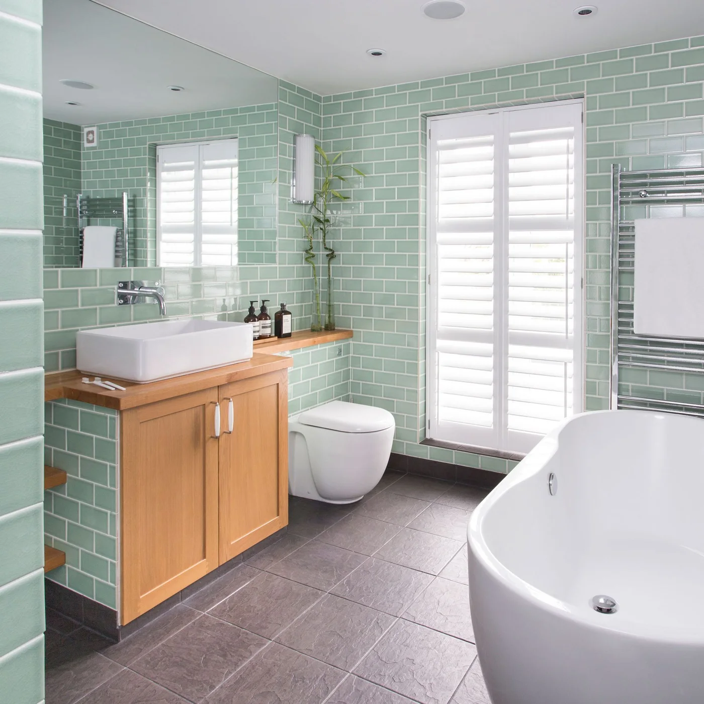 Pale mint-green subway tiles surround a wood vanity, white vessel basin and freestanding bath beside shuttered windows.