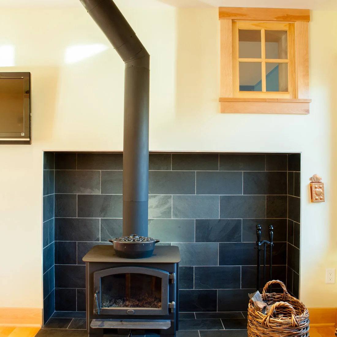 A plain dark stove and tall flue stand within a dark rectangular tiled recess beneath a small wood-trimmed window, with a log basket and fire tools at the side.