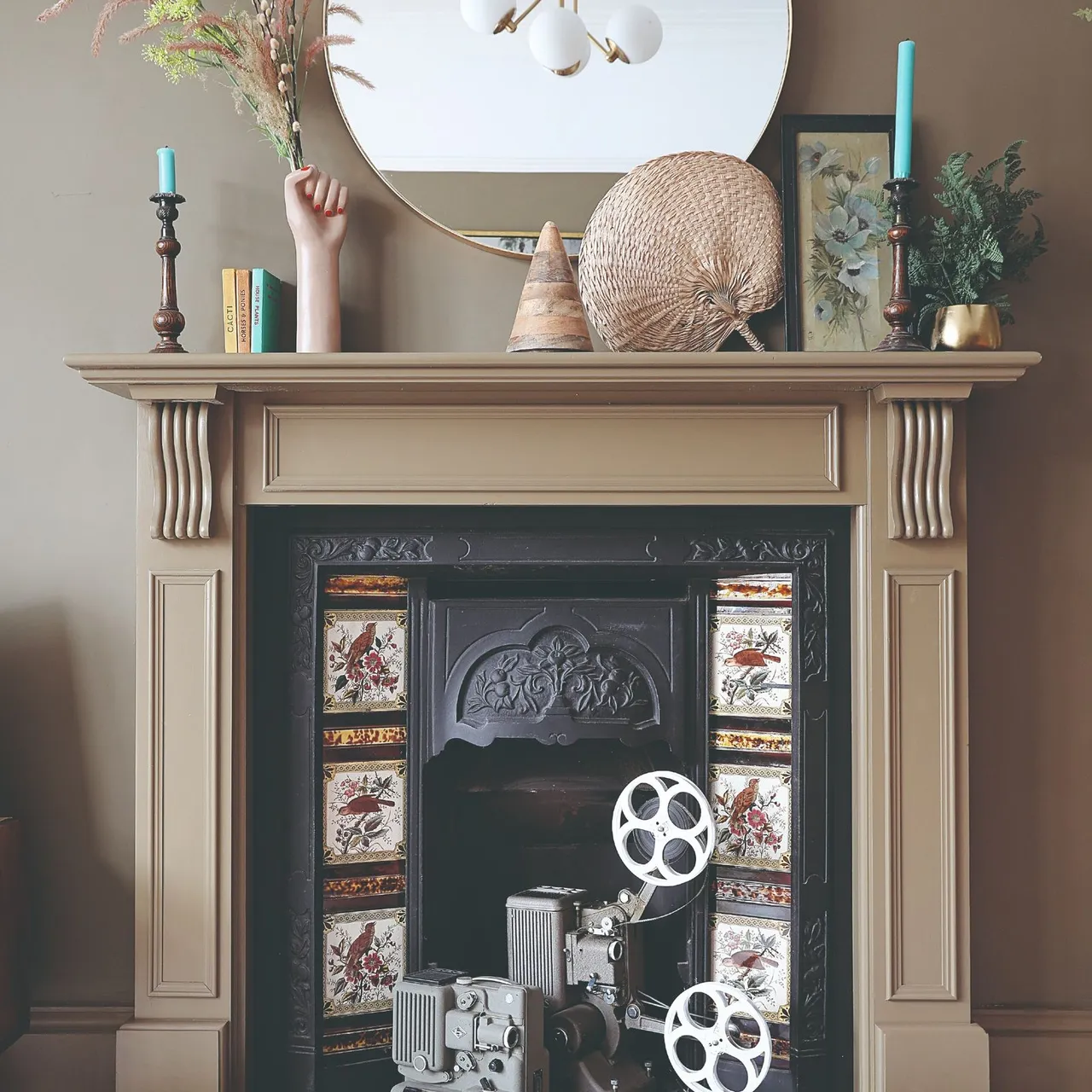 Taupe fireplace mantel with turquoise tapers at its ends, circular mirror, pictures and decorative objects.