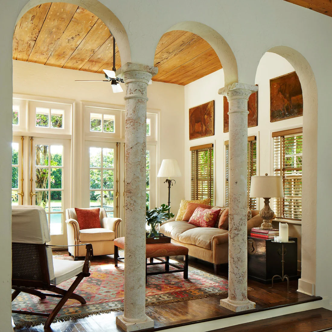 Pale arches on textured columns frame cream living-room seating beneath a wood-toned ceiling beside a warm patterned rug and tall glazed doors
