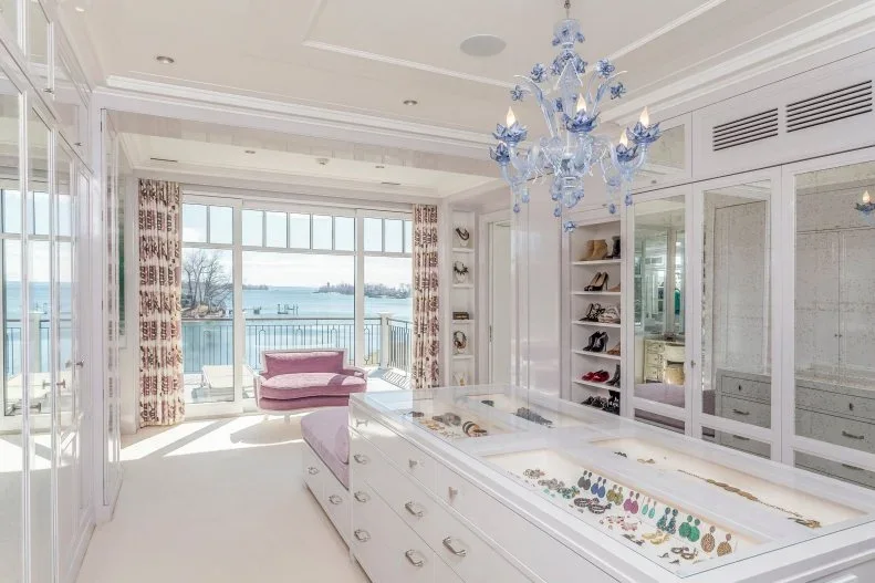 A white closet island displays jewelry beneath a clear top, with mirrored doors and a pink window seat beyond.