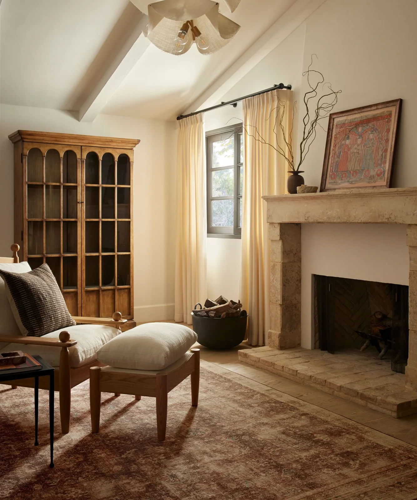 A pale timber-framed chair and footrest sit beside a warm-toned fireplace, tall timber cabinet and cream curtains.