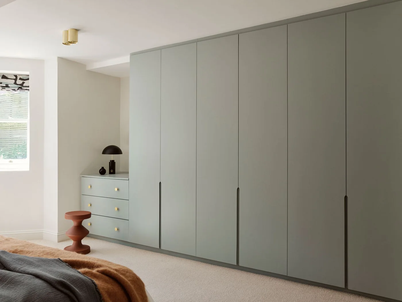 Plain sage-gray wardrobe doors with recessed handles stand beside matching drawers and a rust-colored stool.
