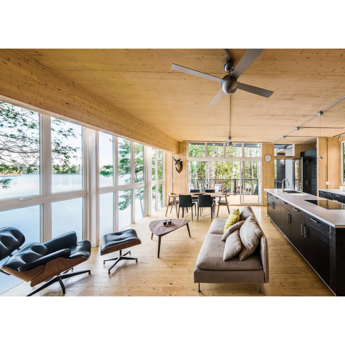 Long wood-lined room with a gray sofa beside a dark kitchen island, black lounge chair and dining table by the far windows.