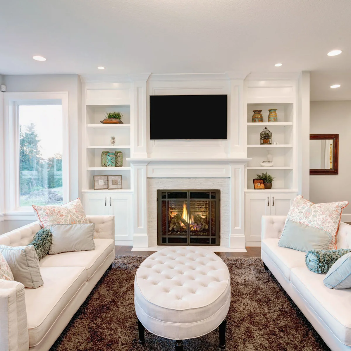 A black television above a white mantel between matching white shelves, with two pale sofas and an oval ottoman.