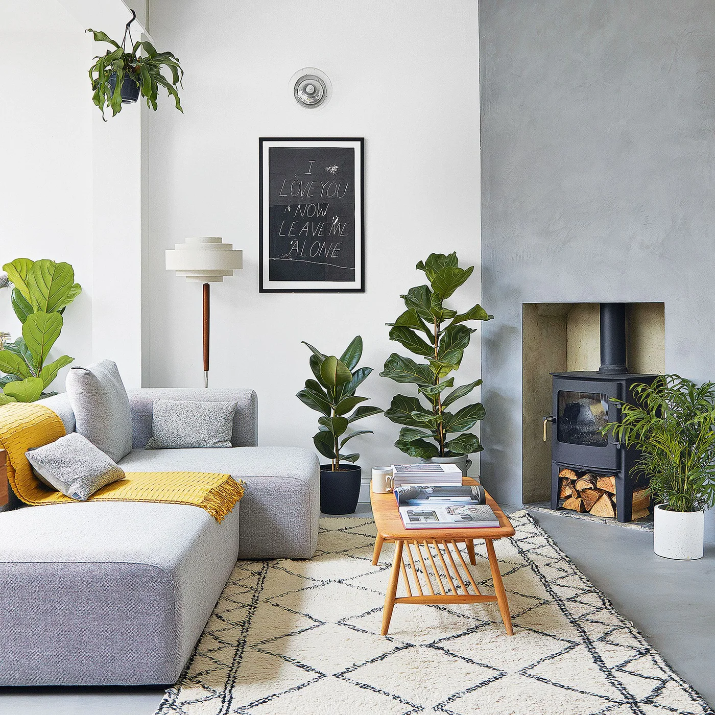 A pale gray chaise sofa with a yellow throw sits near leafy plants and a stove beside a small wooden table on a light patterned rug