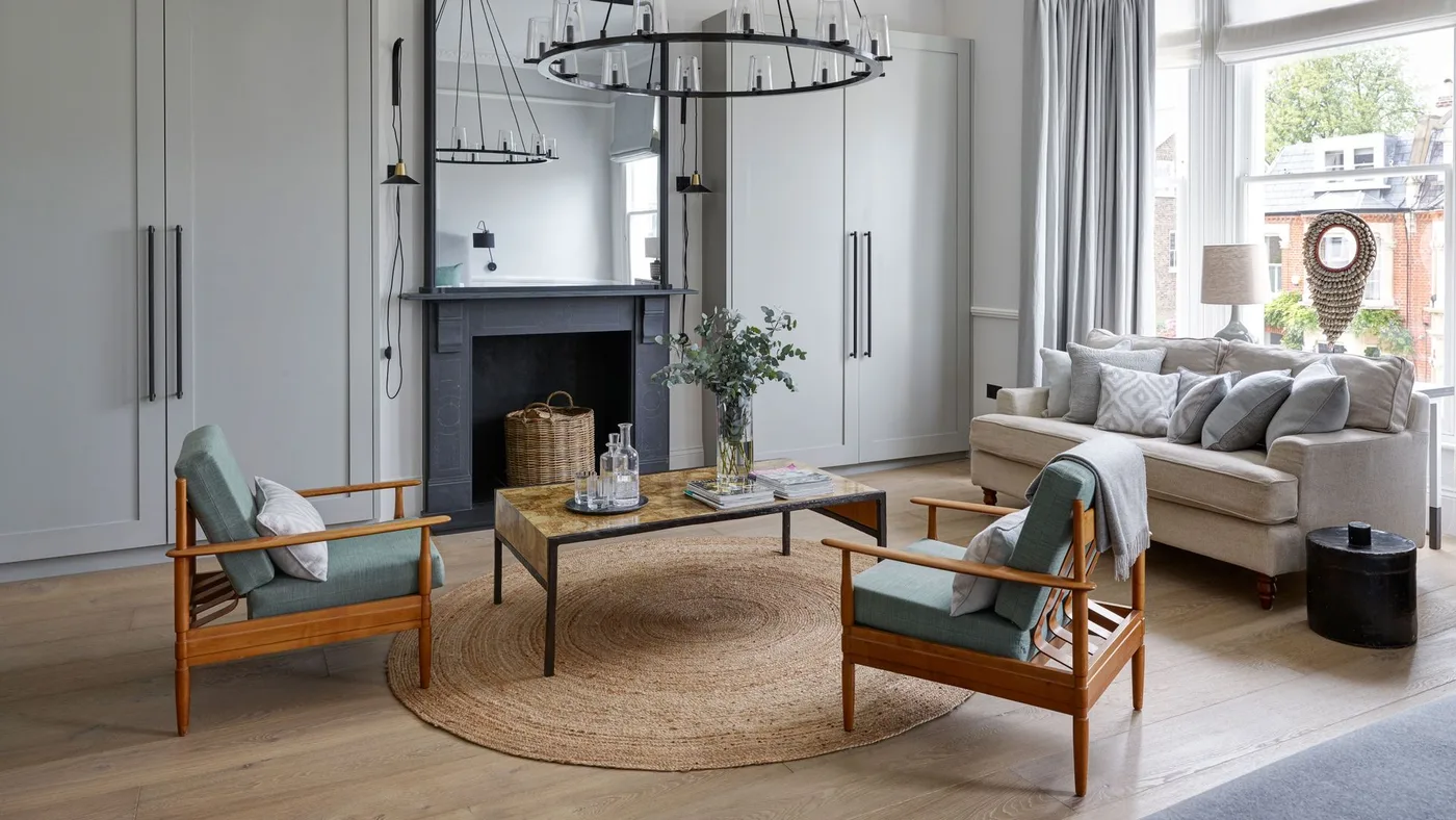 Pale gray living room with full-height cupboards, a light sofa, two wooden-framed chairs and a round woven rug.