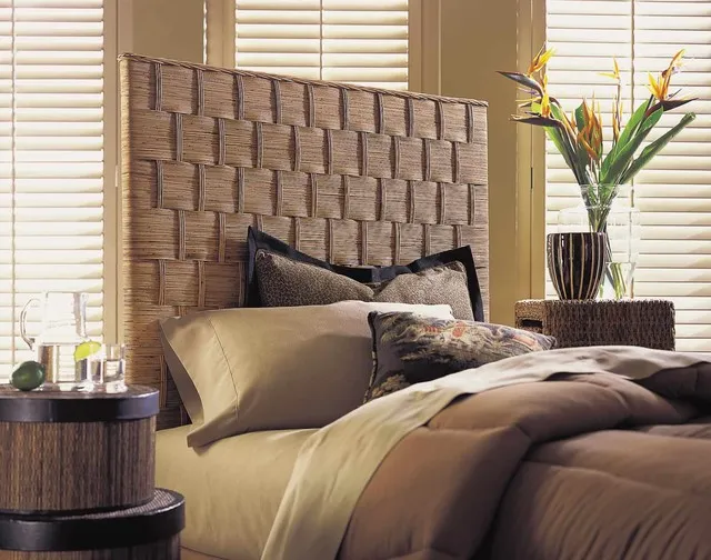 A broad woven headboard sits behind quiet bedding beside a rounded timber bedside table.
