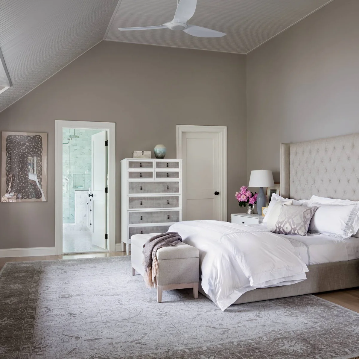 A gray bordered rug extends beyond an upholstered bed and pale bench, with a chest and bathroom doorway nearby.