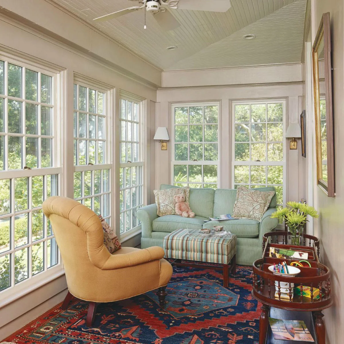 A pale green sofa and yellow armchair face a striped ottoman in a small glazed room with a patterned rug.