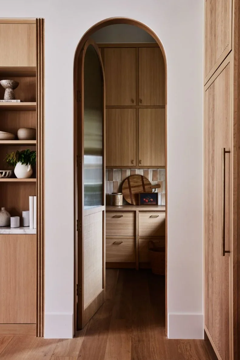 An arched doorway reveals wood pantry cabinets, drawers and a tiled backsplash, with wood shelving and tall cupboards outside.