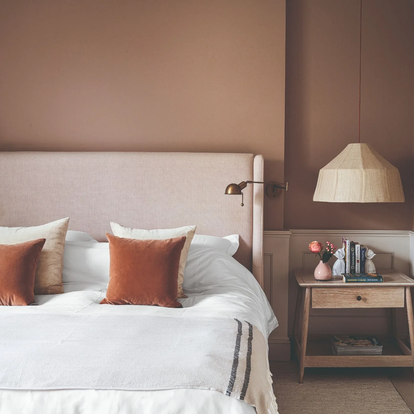 Muted reddish-brown walls with a pale upholstered headboard, rust cushions and a wooden bedside table.