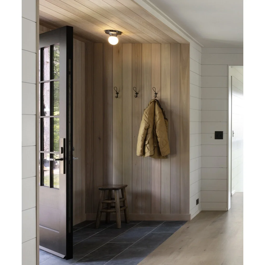 A small timber paneled entry alcove with coat hooks and a stool beside a dark glazed door
