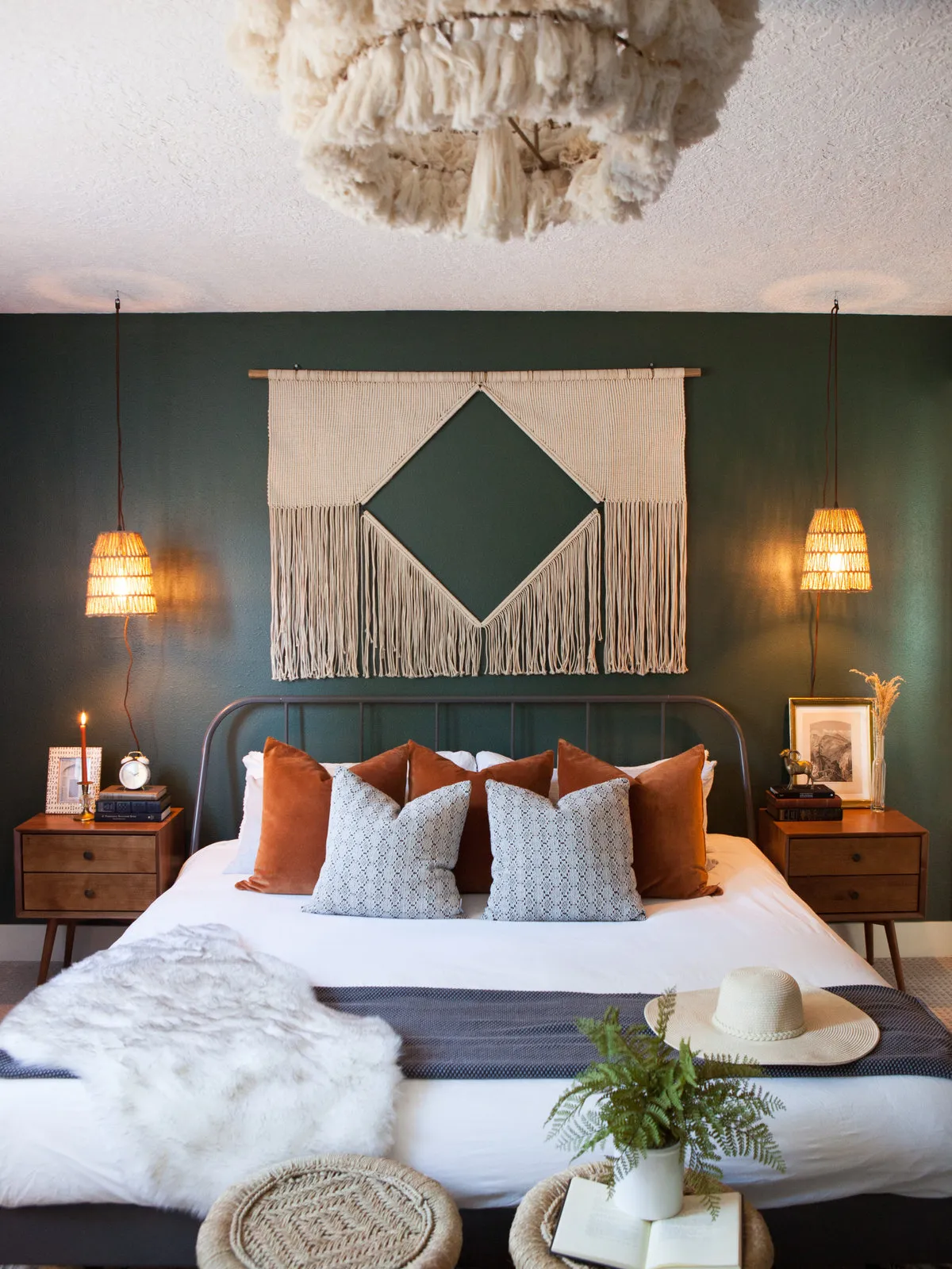 A simple dark metal bed with pale bedding stands against deep green walls beneath a fringed geometric hanging beside wood chests and small woven lights