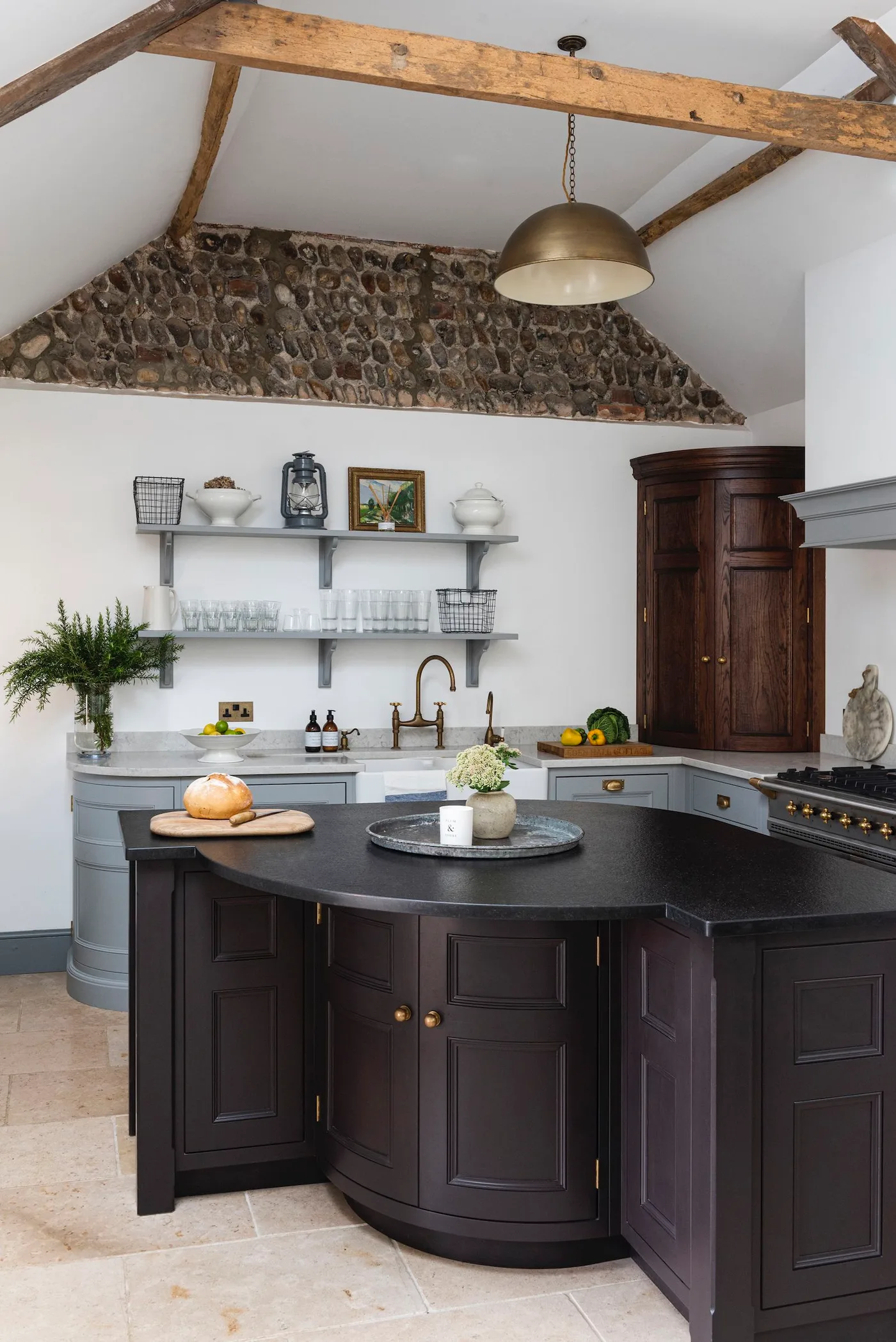 Kitchen with rounded dark island, gray cabinets, exposed beams, rough stone upper wall and large pendant.