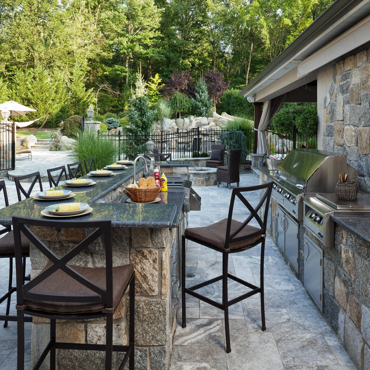 Long dark-topped stone bar with black cross-back stools along its length and around the end beside garden planting