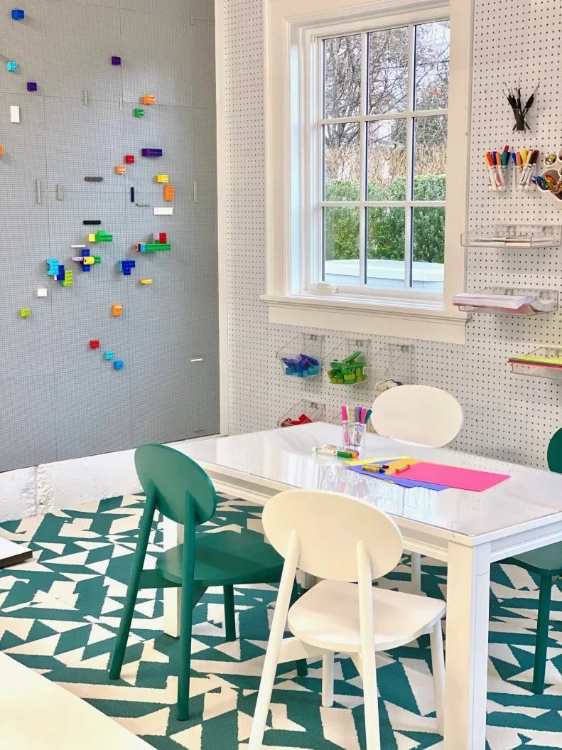A white activity table with green and white chairs stands beside a window, white pegboard tool storage and a gray building-block wall above a green patterned rug.