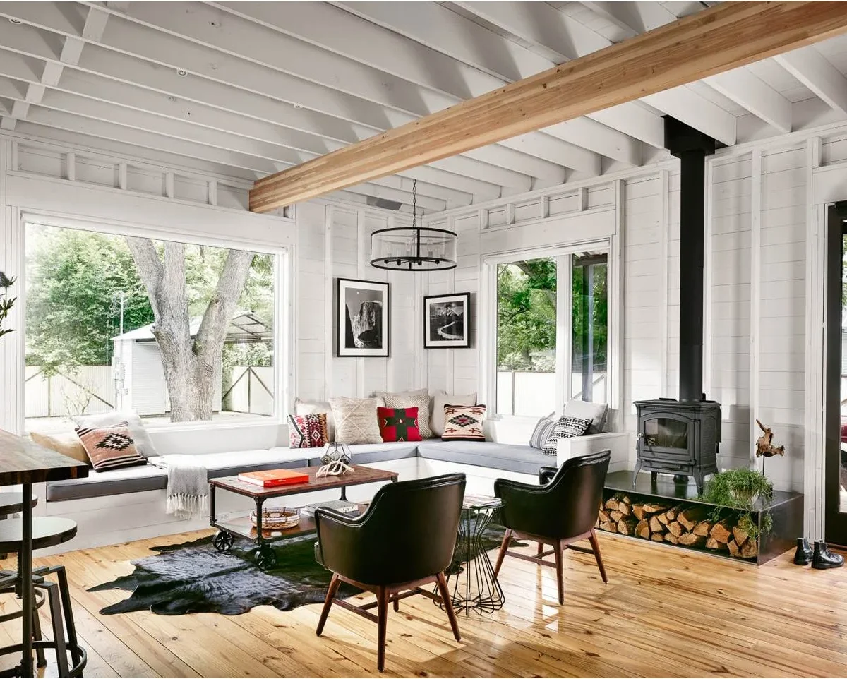 A small black stove and tall pipe sit against pale wall boards in a bright room with built-in window seating and black chairs.