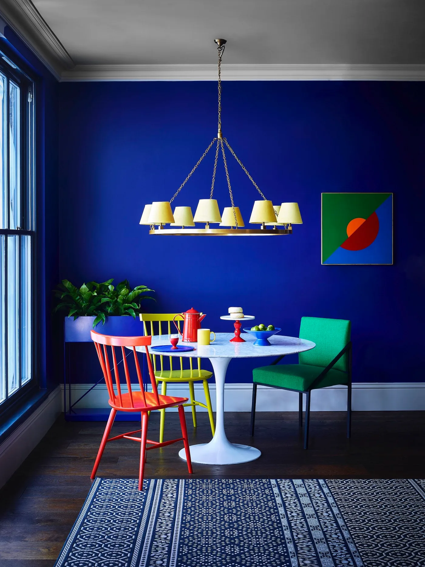 Cobalt blue dining room with a white pedestal table, red and yellow spindle chairs, a green armchair and yellow-shaded chandelier