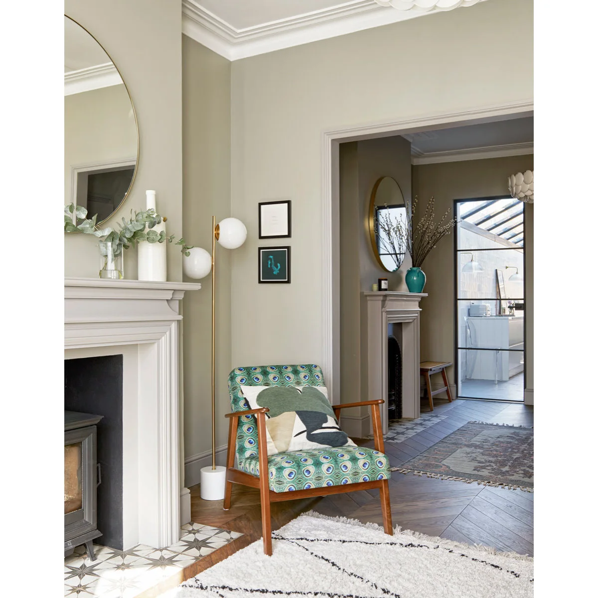 Patterned chair and slim floor lamp beside a pale fireplace with a connected room beyond