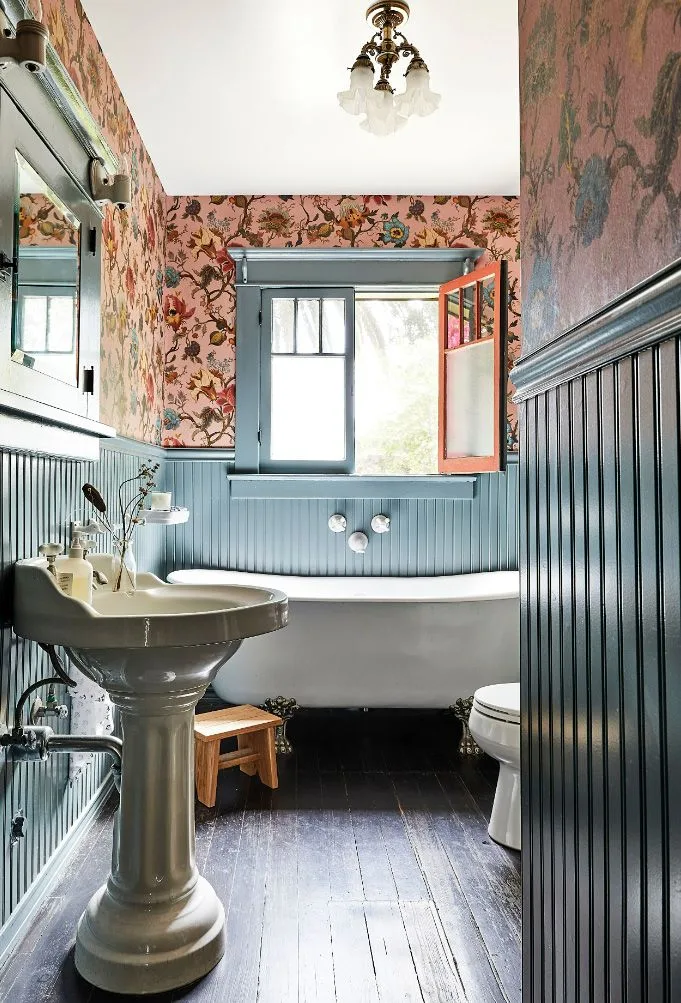 Narrow bathroom with pink floral wallpaper, blue-gray vertical paneling, a pedestal basin, white bath and decorative ceiling light.