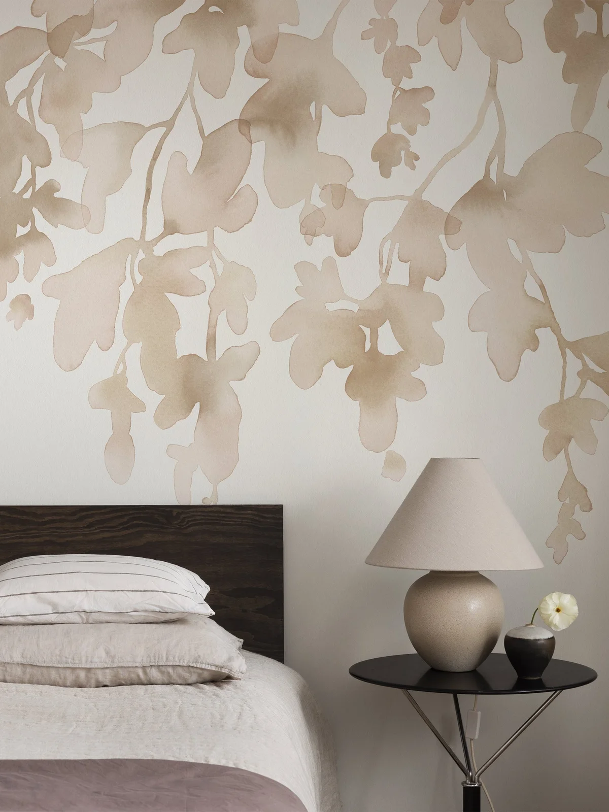 Large beige leaf silhouettes cover a pale wall behind a dark wood headboard, neutral bedding and a round black bedside table with a plain shaded lamp.