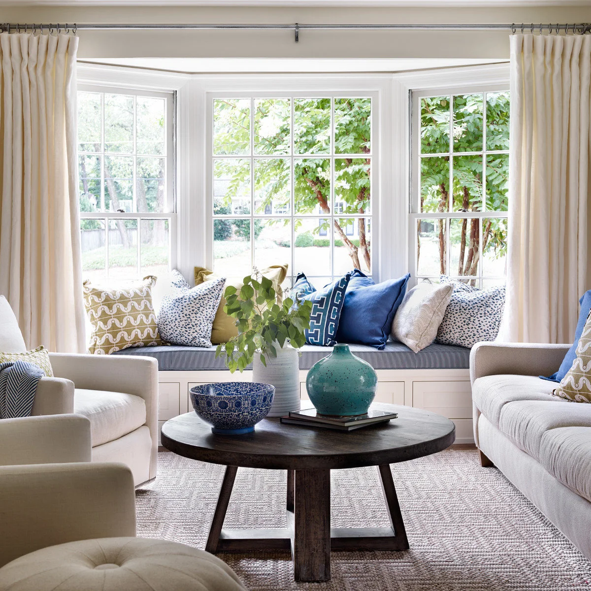 A blue-striped window-seat cushion with blue and green pillows behind a round wood table and pale armchairs.