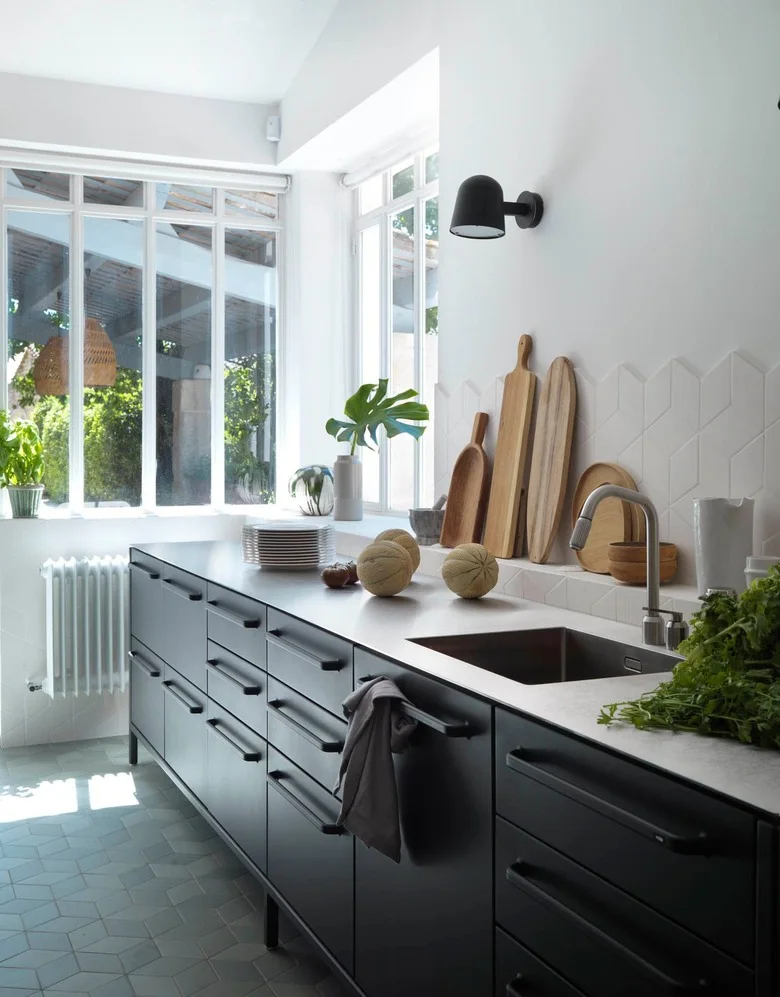 A dark lower cabinet run has wide drawers with straight handles beneath a pale counter, black wall light, white geometric tiles and a broad window.