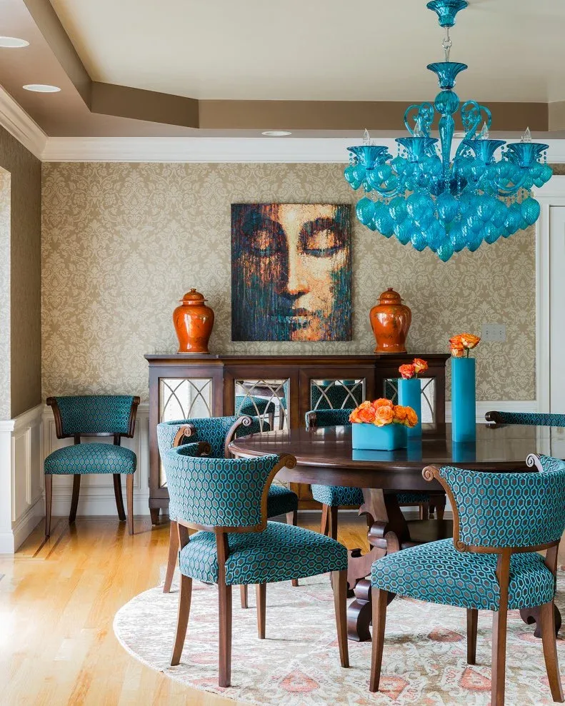 Blue patterned upholstered dining chairs with wooden frames around a dark table, beneath an aqua chandelier, with orange accents on a sideboard.