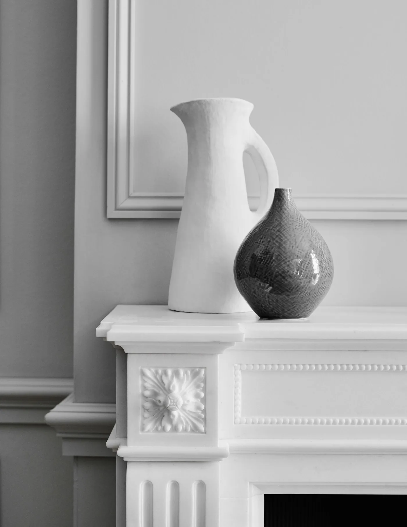 Pale carved mantel with tall white handled jug and smaller rounded dark vase against paneled wall