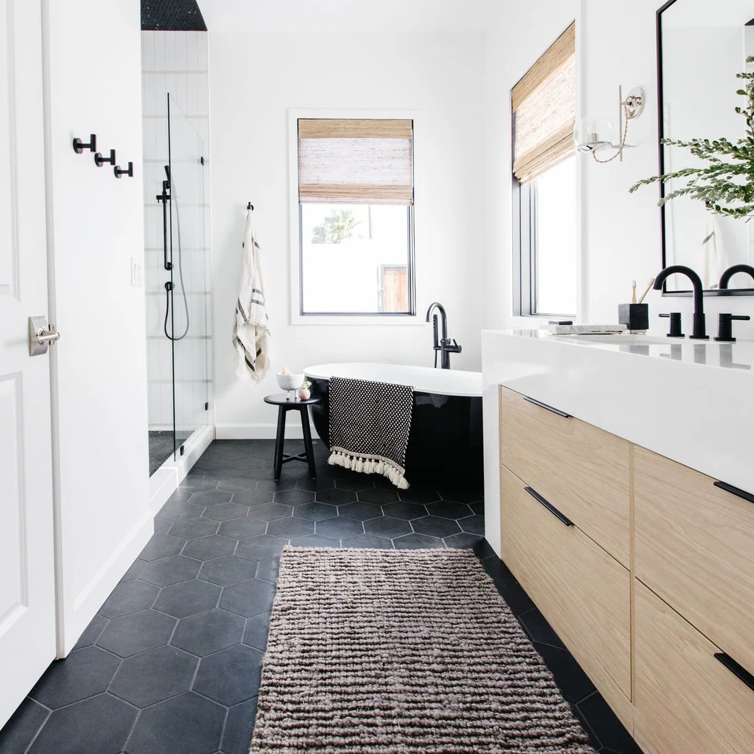 A light wood vanity and black-sided bath stand above dark hexagonal floor tiles beside white walls and woven blinds.
