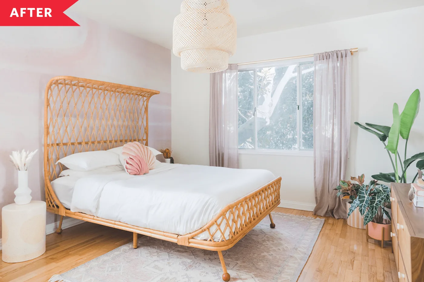 Bright bedroom with woven bed frame, white bedding, pink shell-shaped pillow, pale curtains, woven pendant light and broad-leaved plants beside the window.
