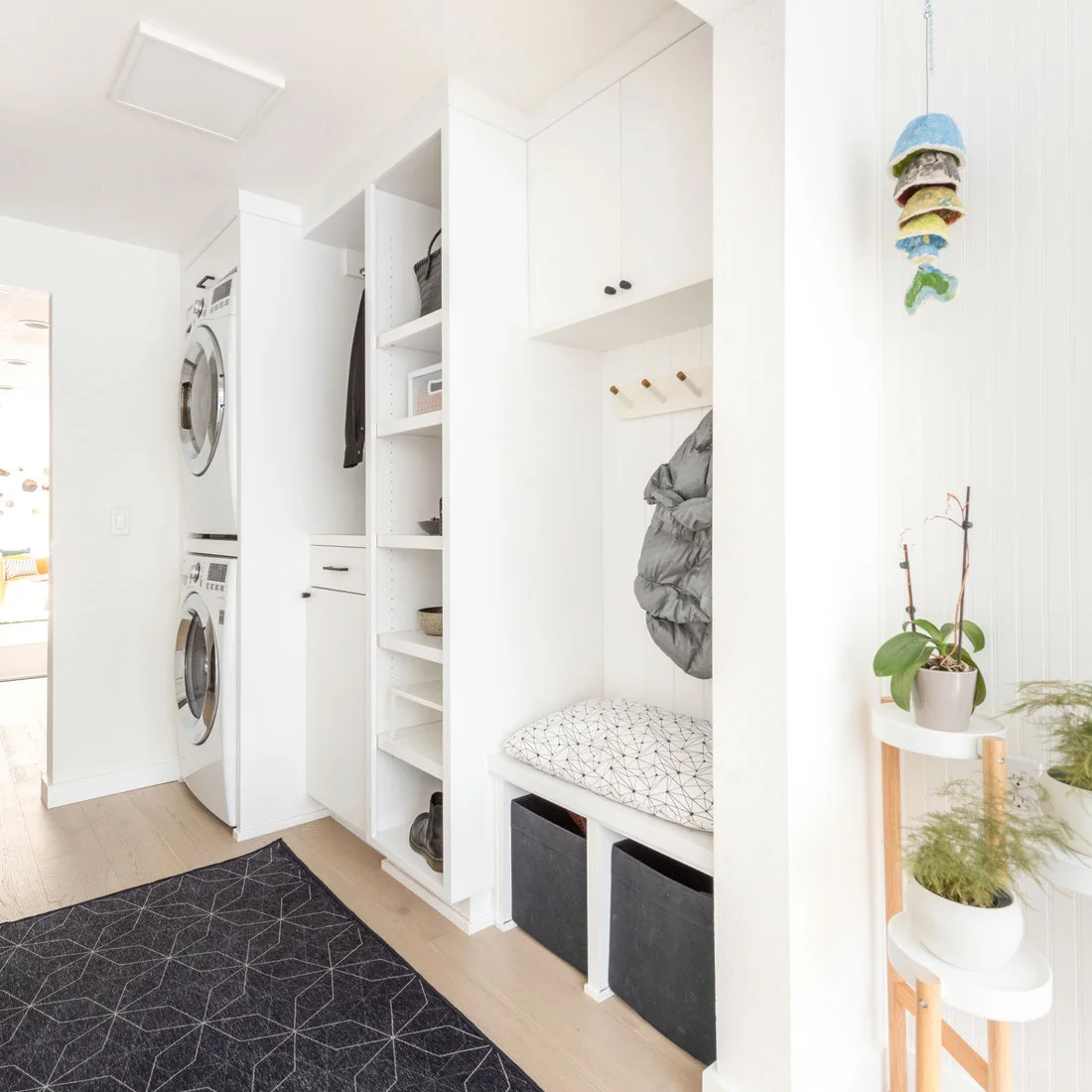 Stacked white laundry machines stand beside tall white cubbies, a cushioned bench and wood coat pegs on a pale wood floor.