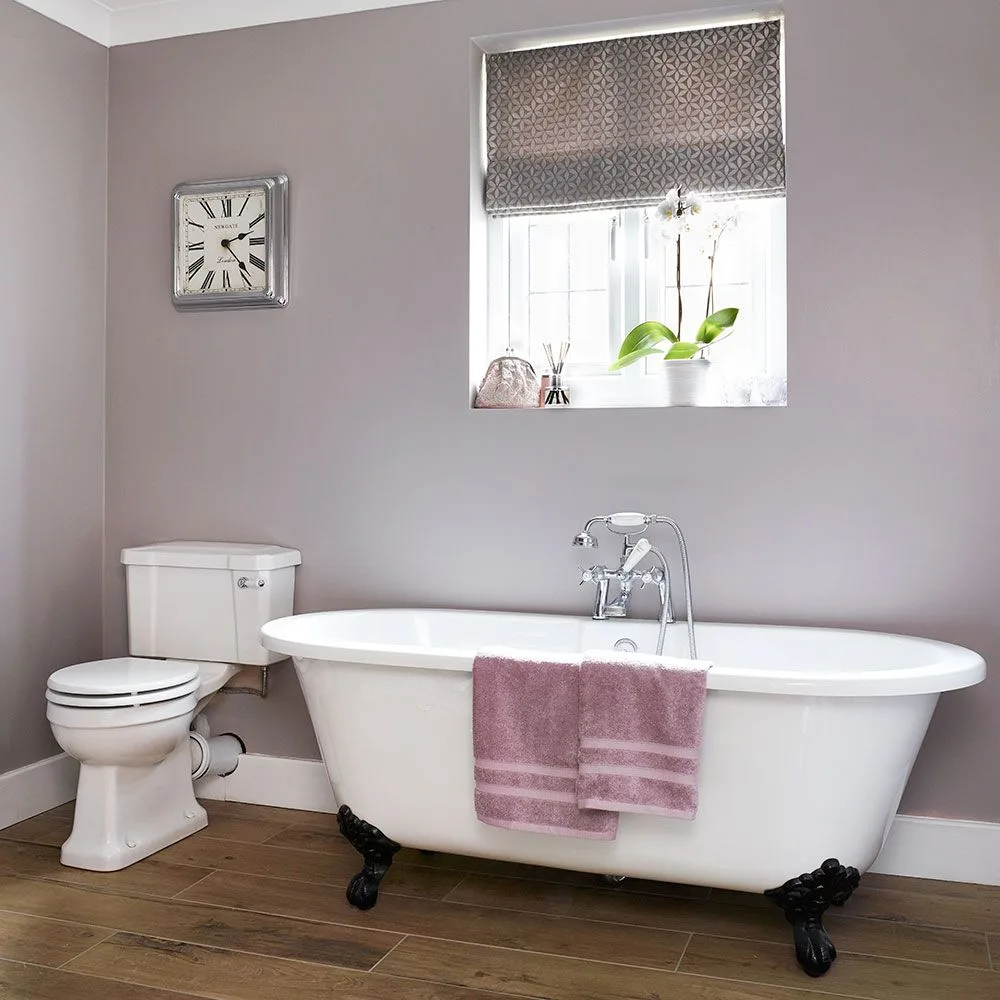 White claw-foot tub against muted lavender-gray walls, with a patterned blind and wood-toned floor.