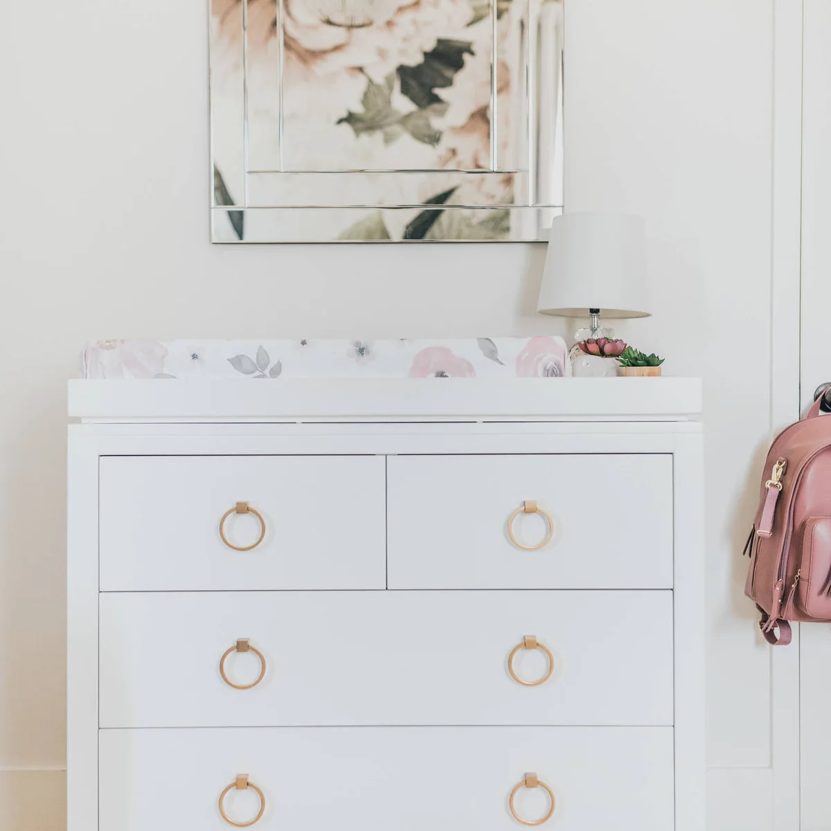 White nursery changing chest with gold-colored ring pulls, a floral pad and a framed mirror