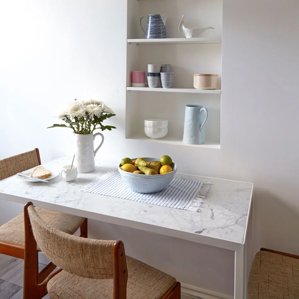 A pale rectangular dining table and woven chairs sit beside a recessed shelf filled with cups, bowls and jugs.