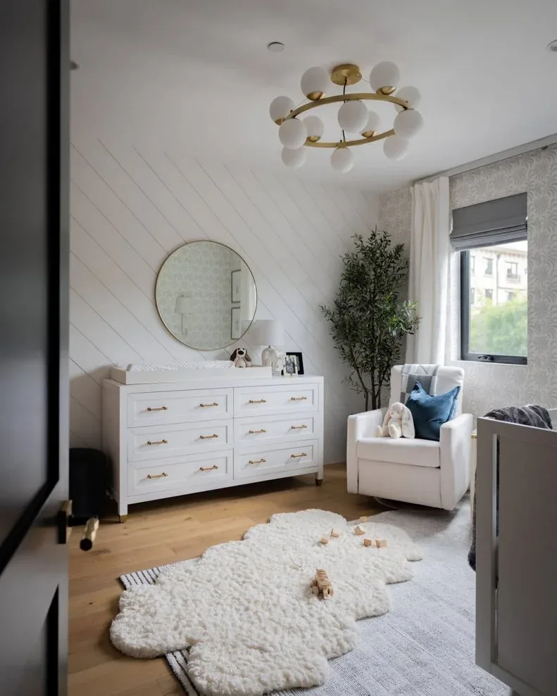 Nursery with diagonal white board wall, white dresser, round mirror, pale chair, wood floor and layered rugs
