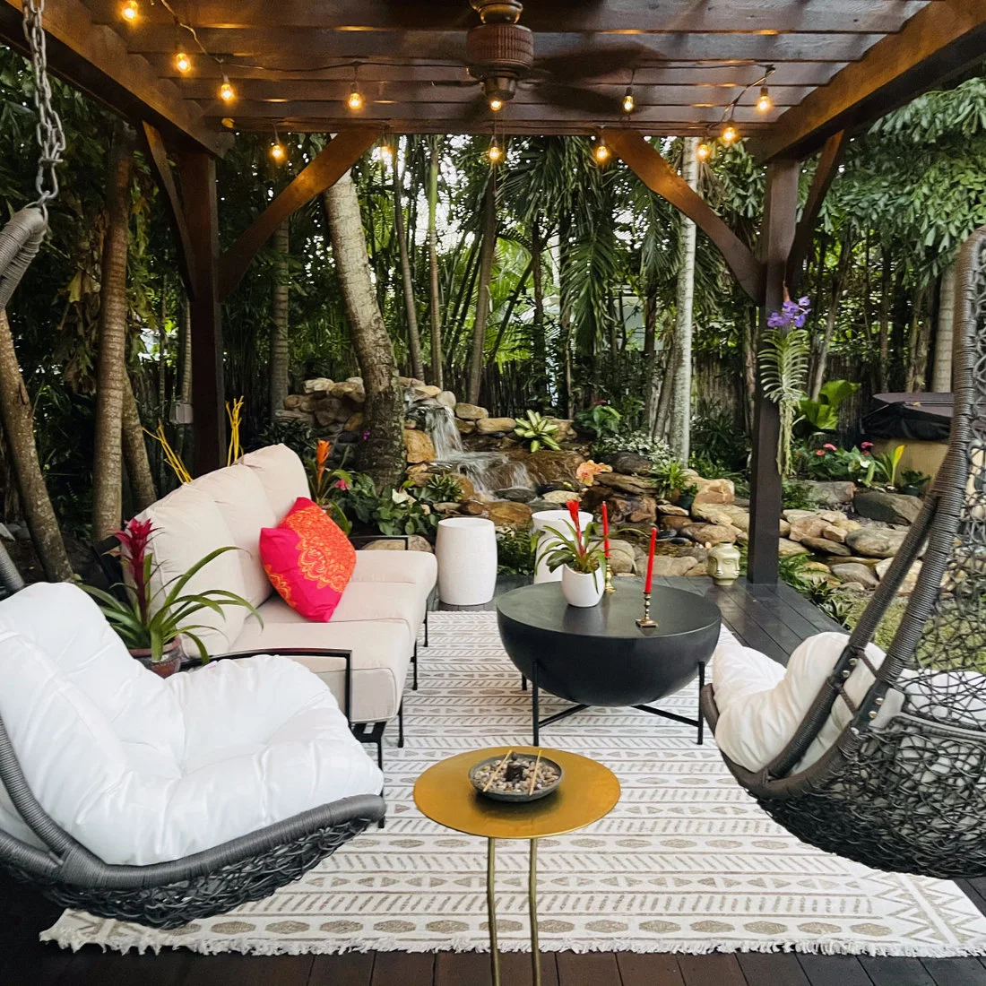 Dense palms and leafy planting surround pale cushioned seating beneath a timber garden roof