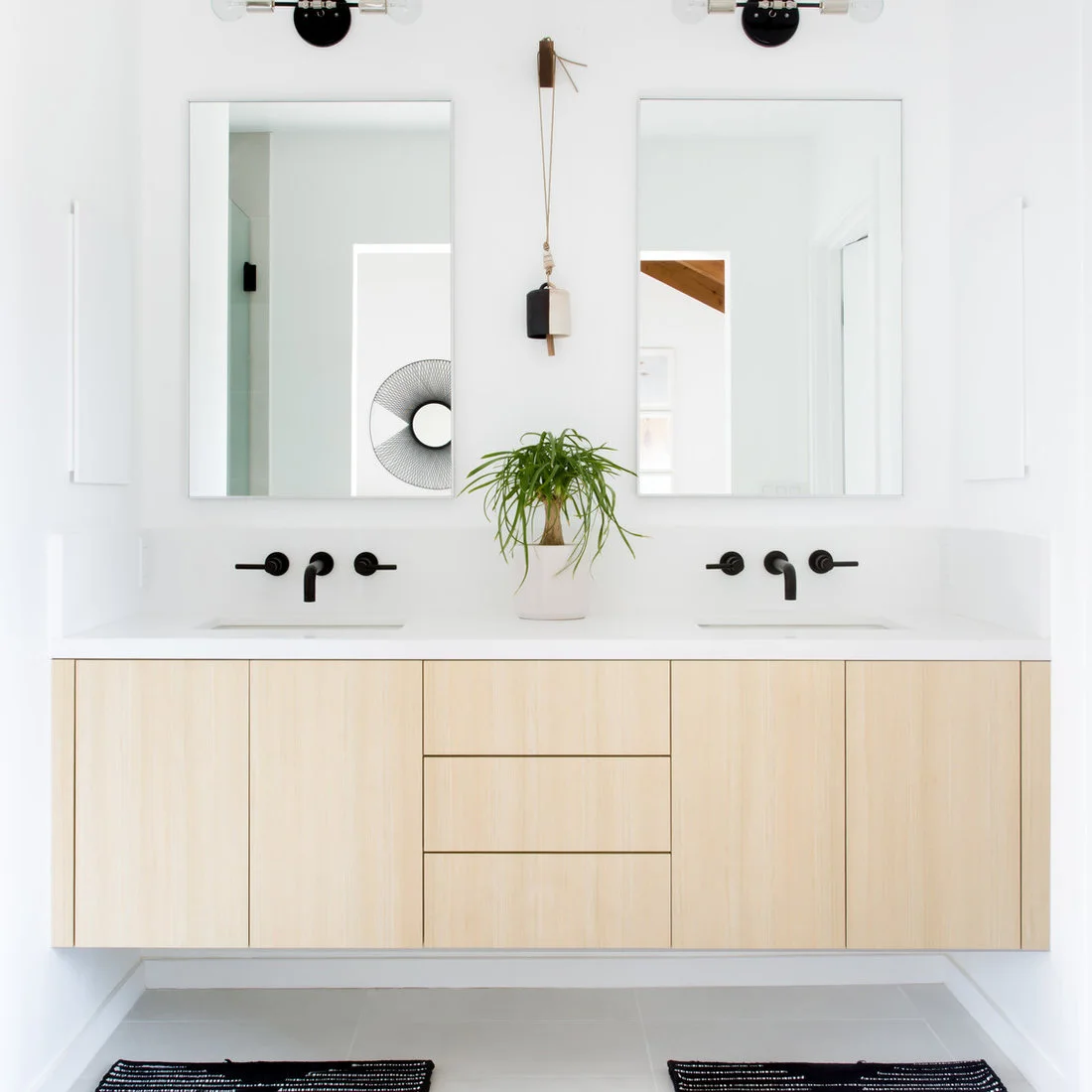 A wall-hung pale wood double vanity sits beneath two rectangular mirrors and black wall taps, with a small plant between the basins and striped rugs below.