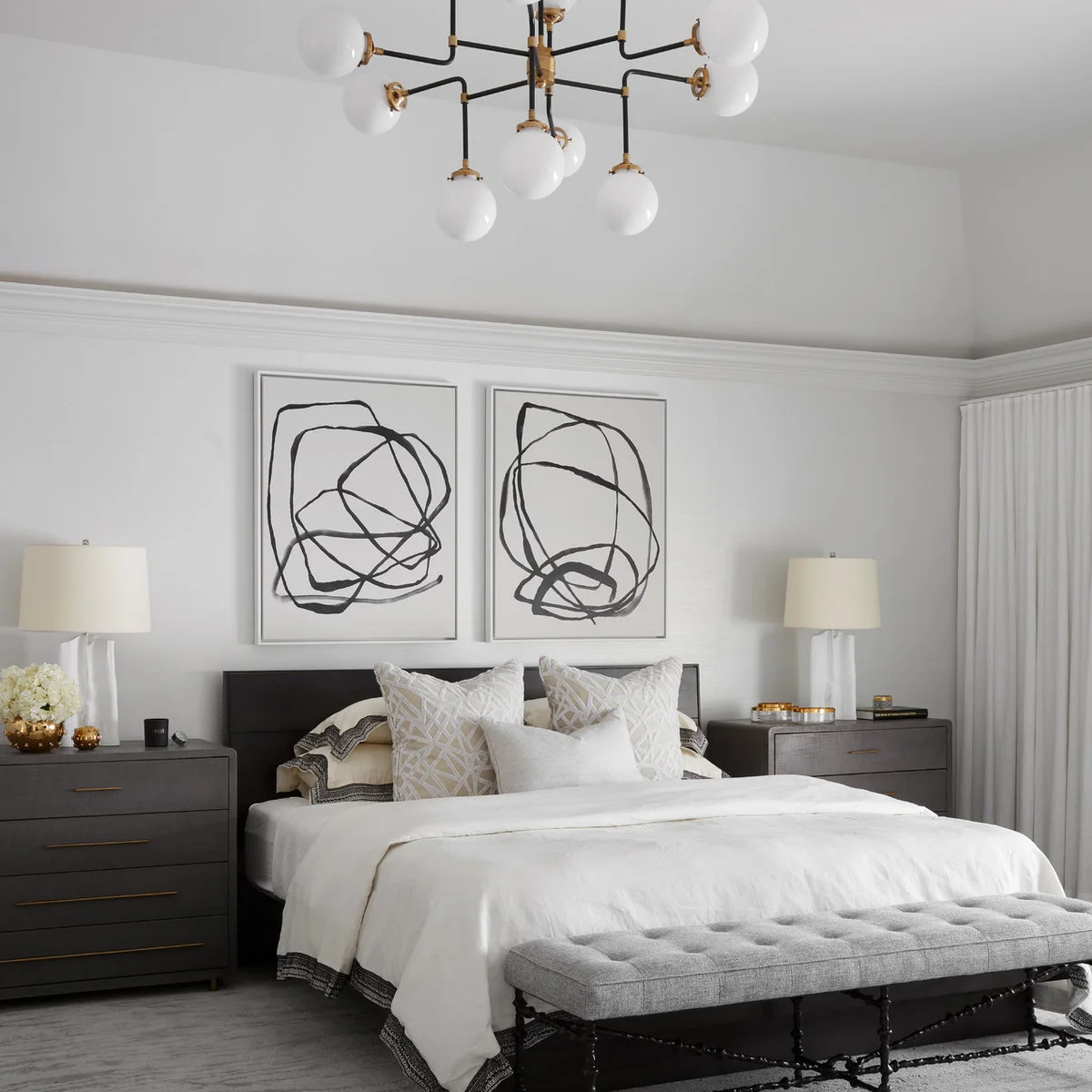 Wide dark bedside chests flank a pale bed beneath two abstract drawings, with a long gray bench at the foot.