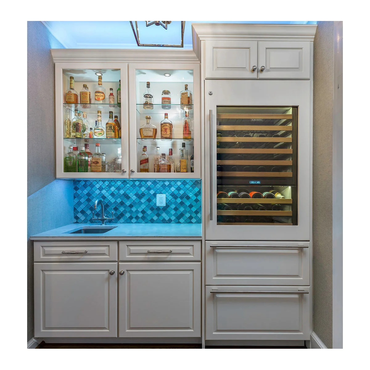 White mini bar with blue mosaic backsplash, small sink, illuminated bottle cupboard and tall wine refrigerator