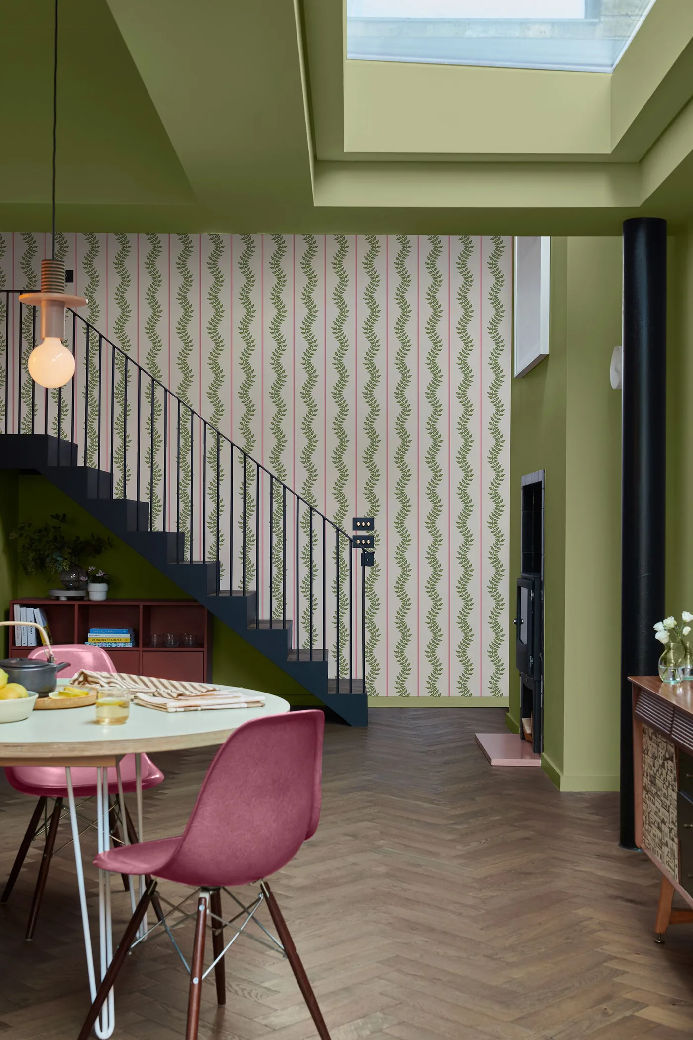 Green dining area with leafy striped wallpaper beside a dark staircase, pale round table and pink molded chairs