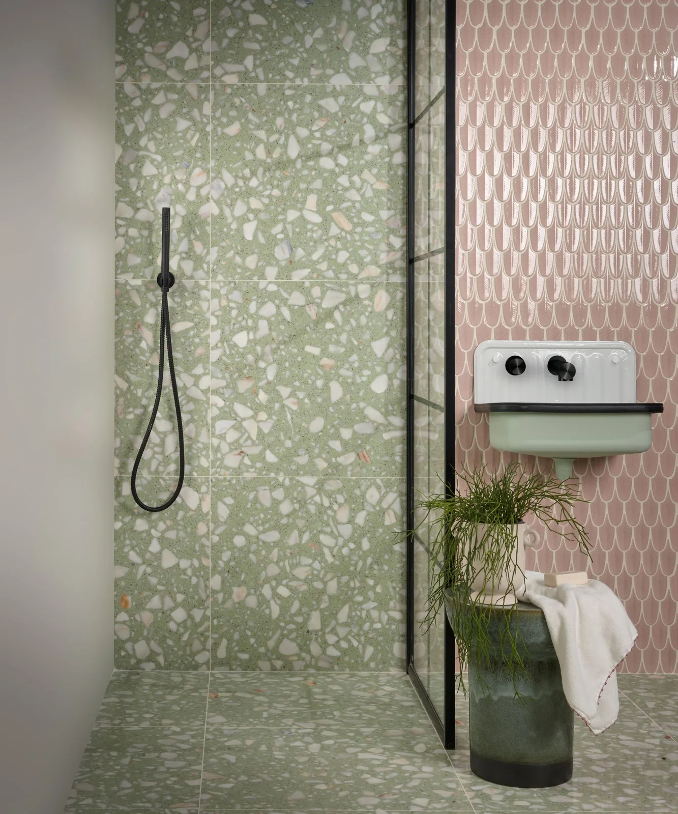 Green terrazzo surrounds a shower beside pink scallop tiles and a small pale green wall-mounted basin.