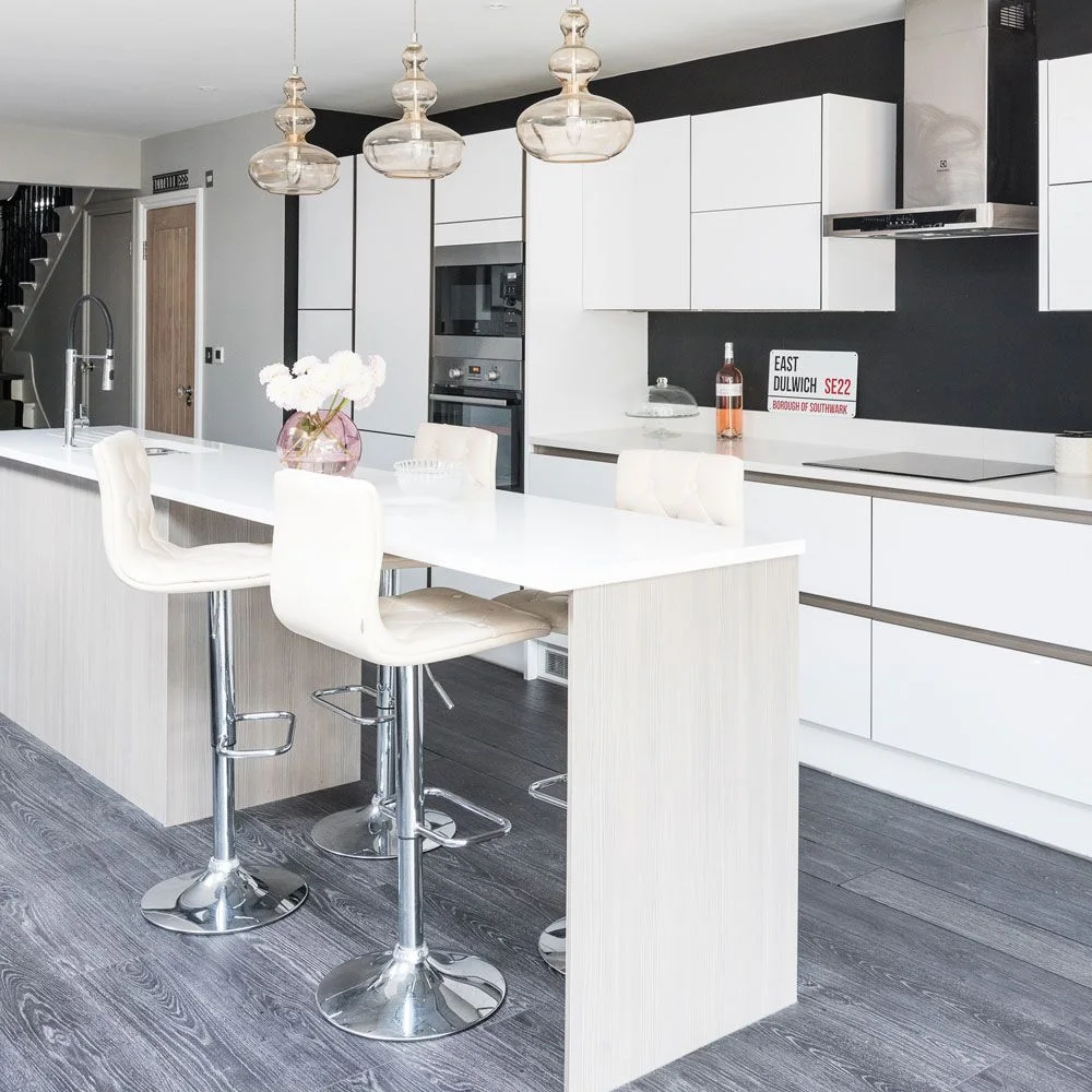 Flat white cabinet doors and wide drawers stand against a black backsplash wall beside a pale wood-look island.