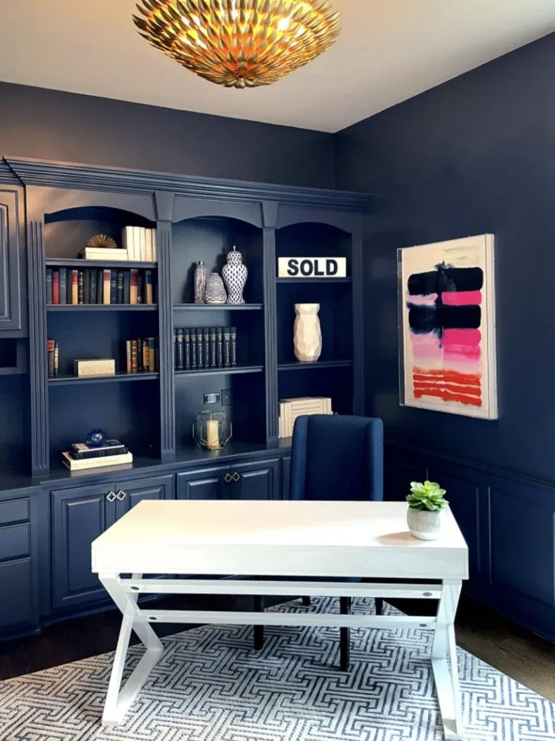 Deep blue walls and matching bookcases surround a pale desk and blue chair beneath a sculptural warm-colored ceiling light.