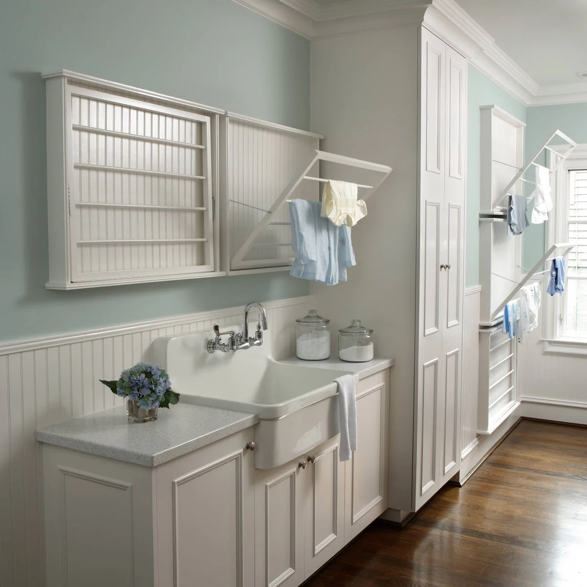 Pale blue laundry room with white fold-down wall drying racks, a large apron-front sink and tall cabinets