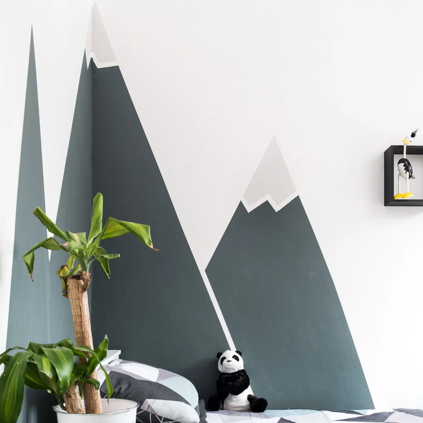 A large dark and pale mountain mural stands behind a child’s bed with geometric bedding, a panda toy and a small dark wall box