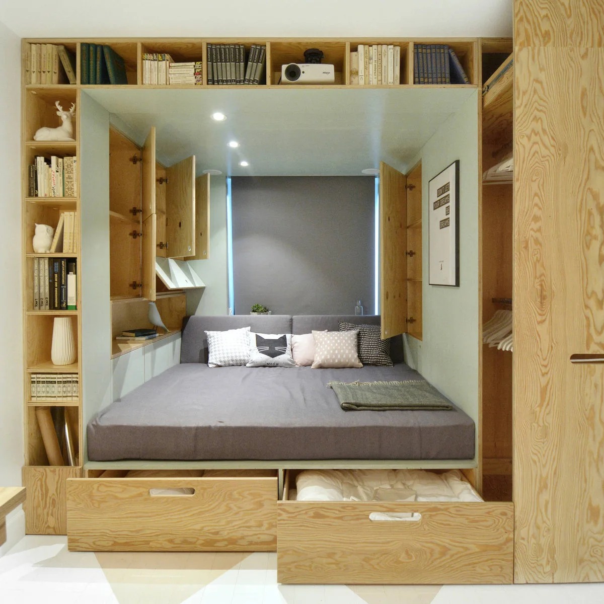 Compact sleeping alcove with open drawers beneath a gray bed and wood shelves around the opening
